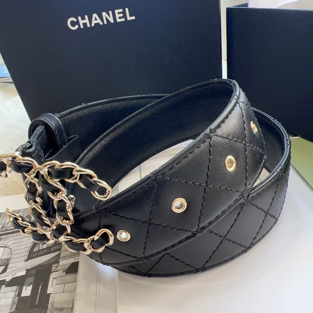 Black quilted leather belt with gold chain detail on a white surface, with Chanel branding in the background.