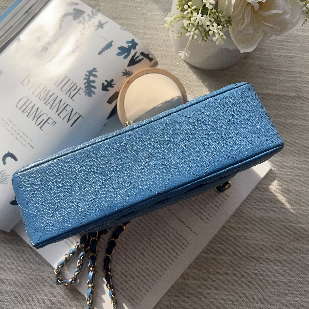 Blue quilted handbag on a wooden surface with a book and flowers in the background