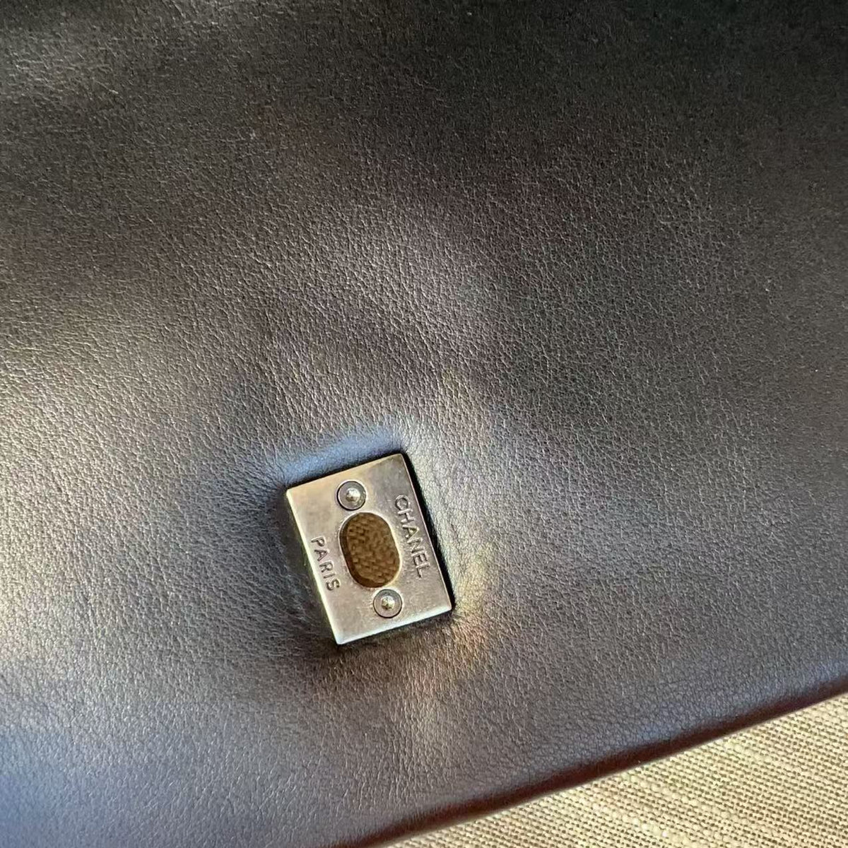 A close-up of a small silver-toned metal square hardware piece with a circular gold center, placed on a dark brown textured leather surface with a beige woven fabric visible in the lower right corner.