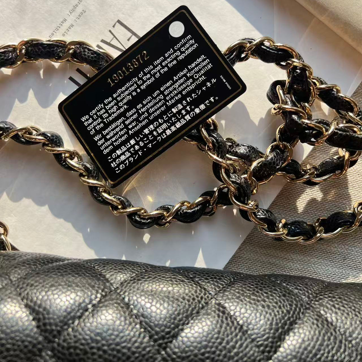 Close-up of a black quilted leather handbag strap with a gold-tone chain link and a black rectangular metal nameplate, placed on white paper packaging.