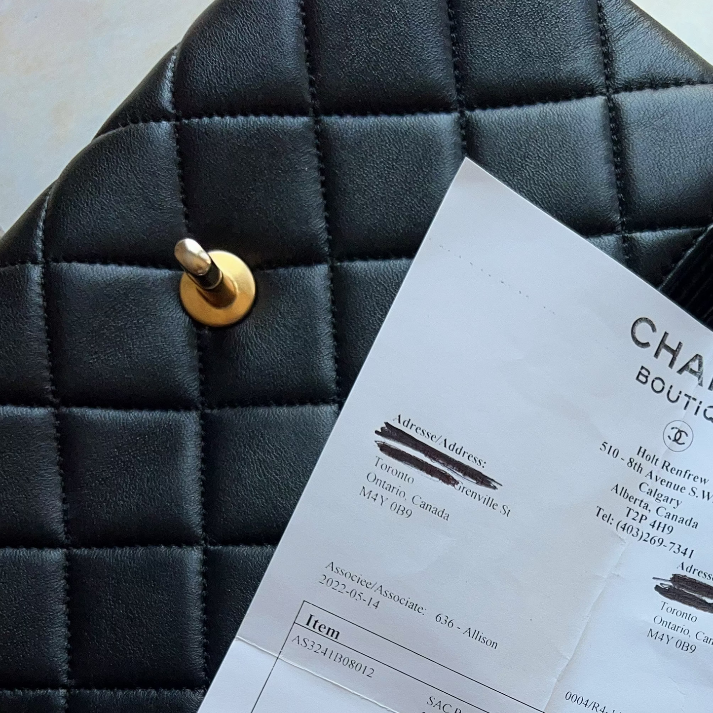 A black quilted Chanel handbag with gold-tone hardware lies on a light beige fabric surface, with a branded Chanel Boutique receipt sheet placed on top.