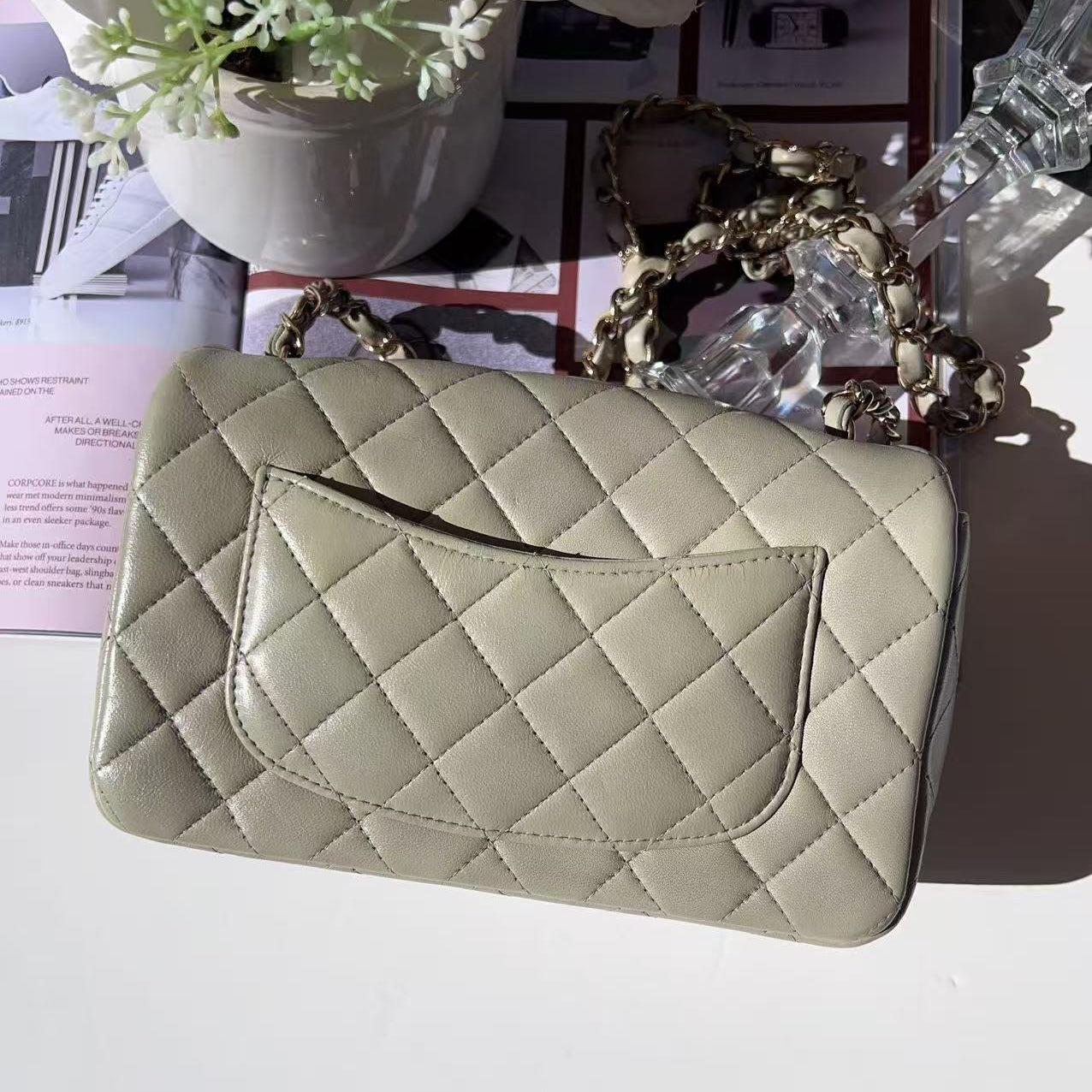 Quilted beige handbag on a white surface with a magazine and decorative items in the background