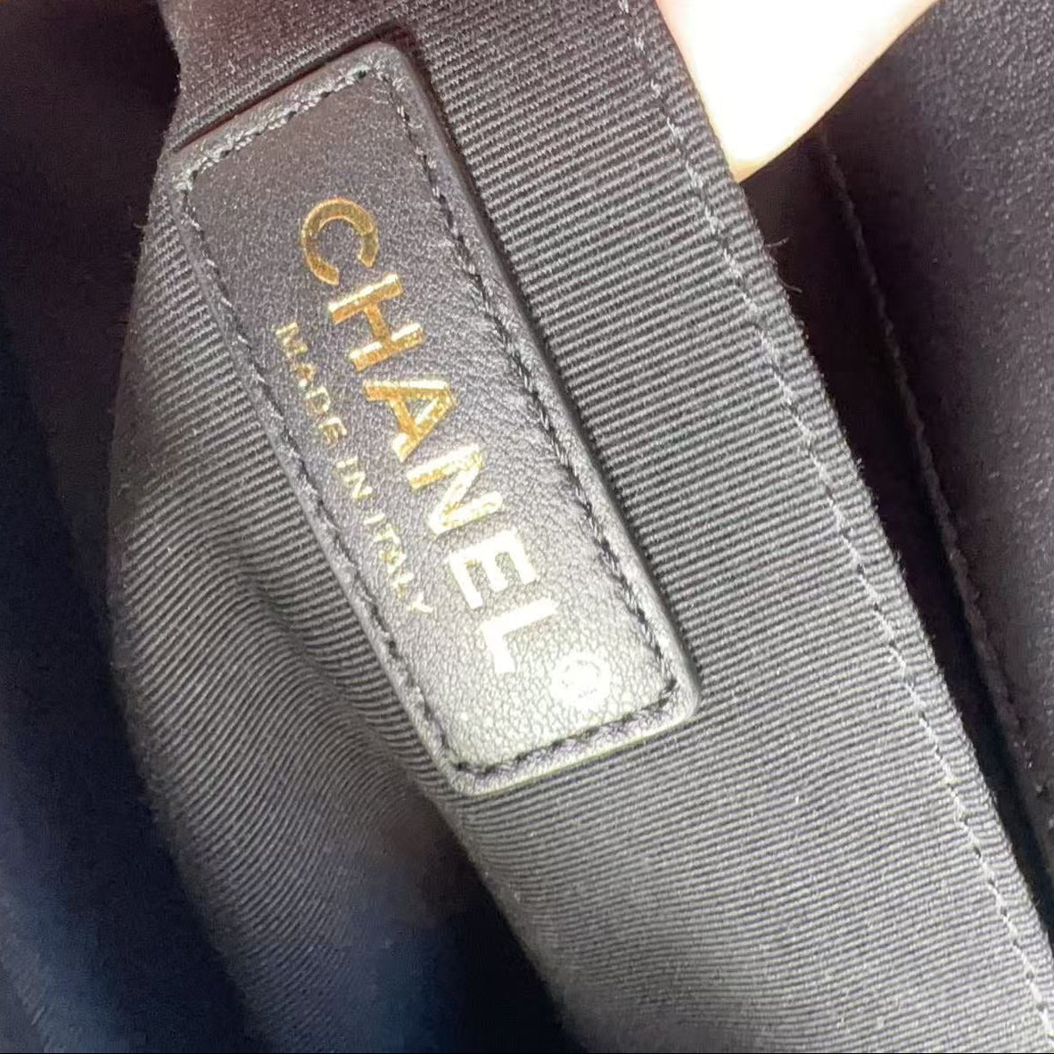 Close-up of a black Chanel interior label with gold lettering, held by fingers against a black fabric background.