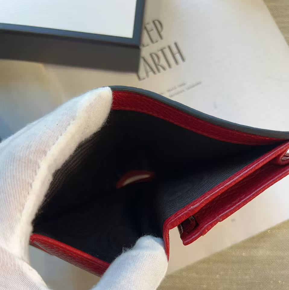A close-up of a red leather wallet or card holder being held open by a white-gloved hand, with a dark interior lining and part of a branded box visible in the background.