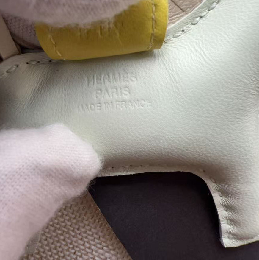 Close-up of a Hermès Paris leather shoe detail, showing the embossed brand name on a pale green leather piece with a yellow pull tab, held by white-gloved hands.