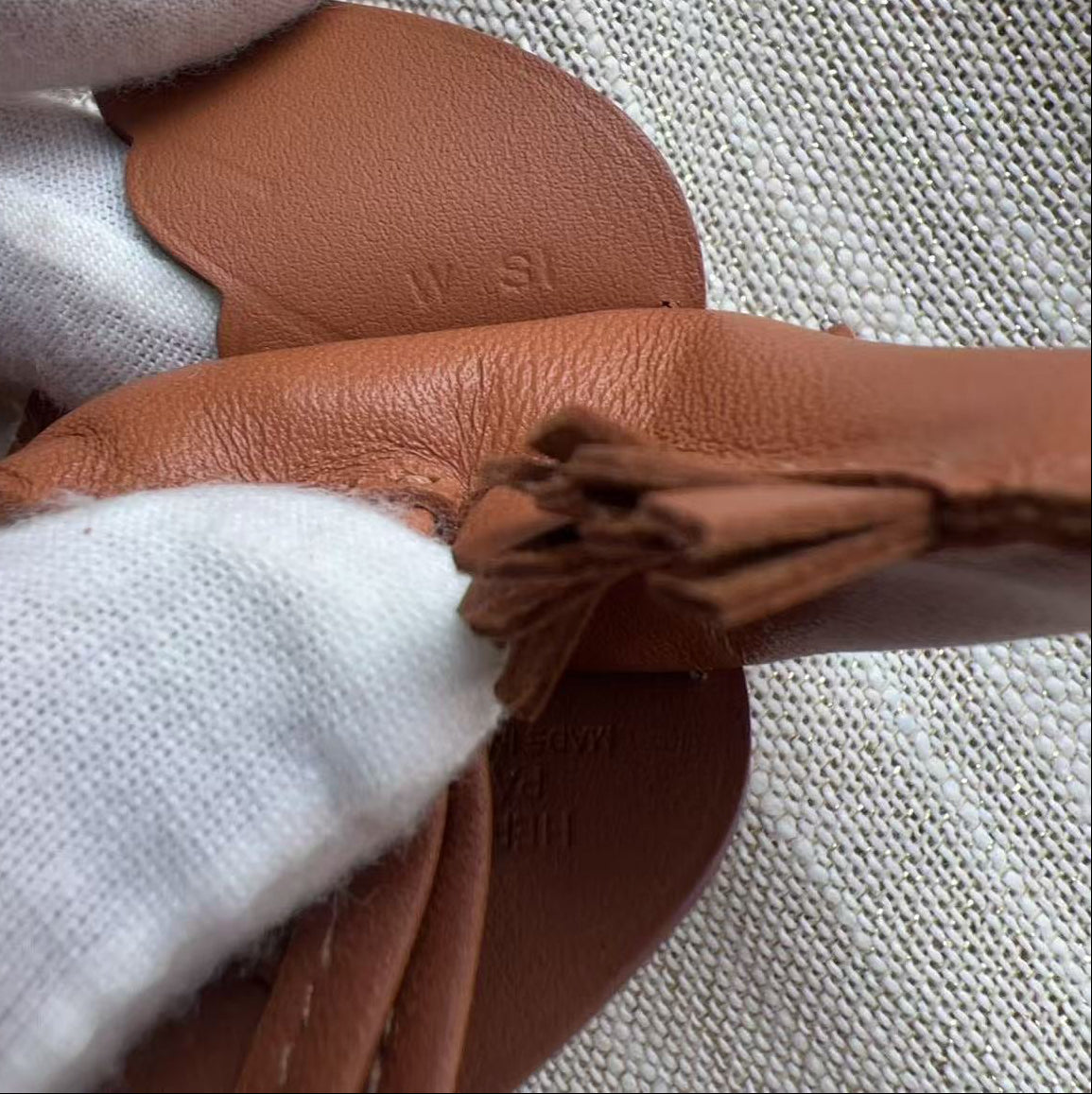A close-up of a person wearing white knit gloves holding a tan leather strap or accessory against a textured beige fabric background.