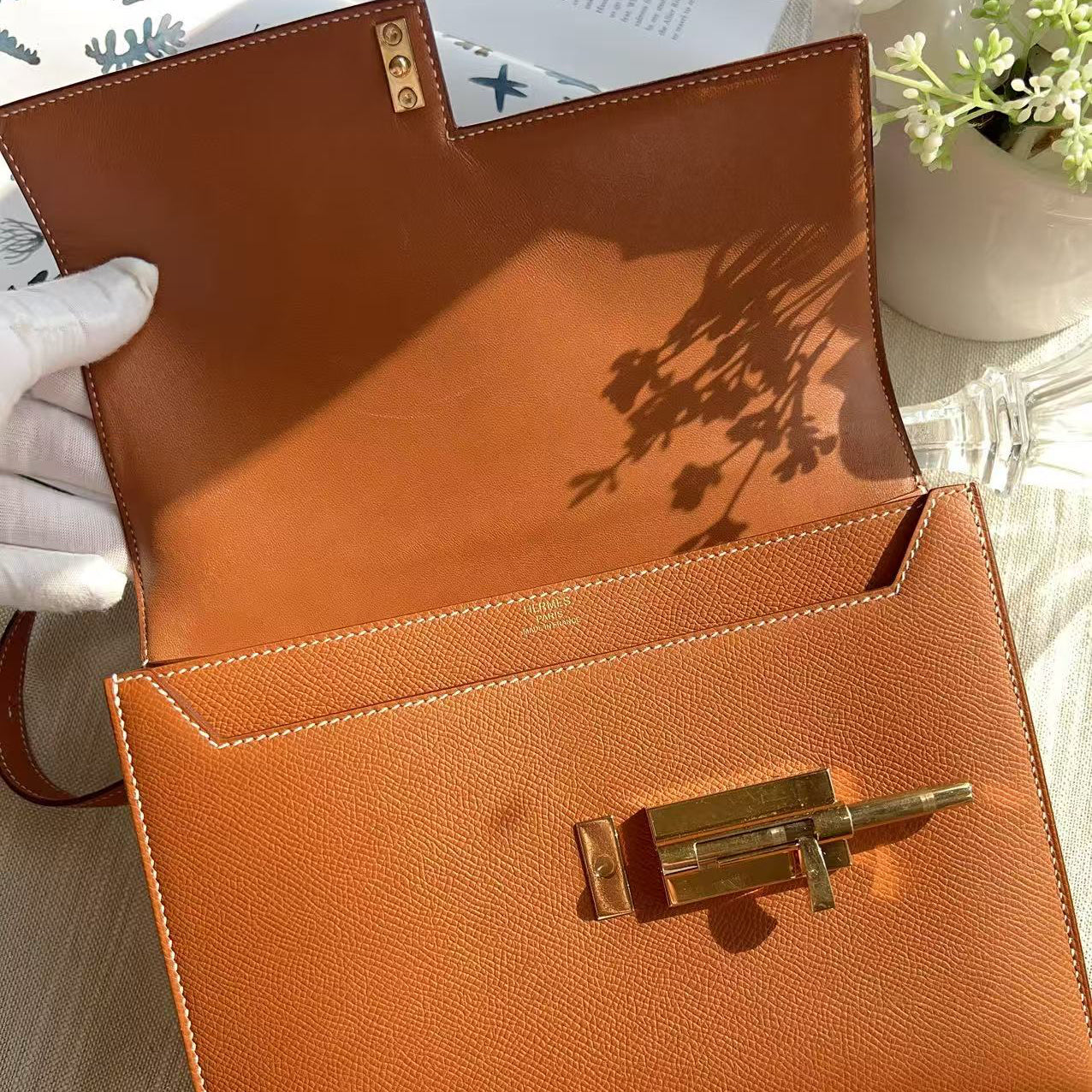 Brown leather clutch with gold clasp held by a gloved hand on a light wooden surface.