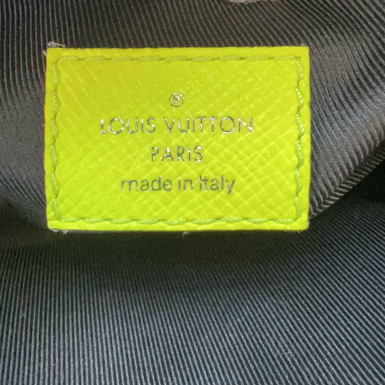 Close-up of a Louis Vuitton Paris made in Italy interior label sewn onto dark gray fabric, with a white gloved finger visible at the top.