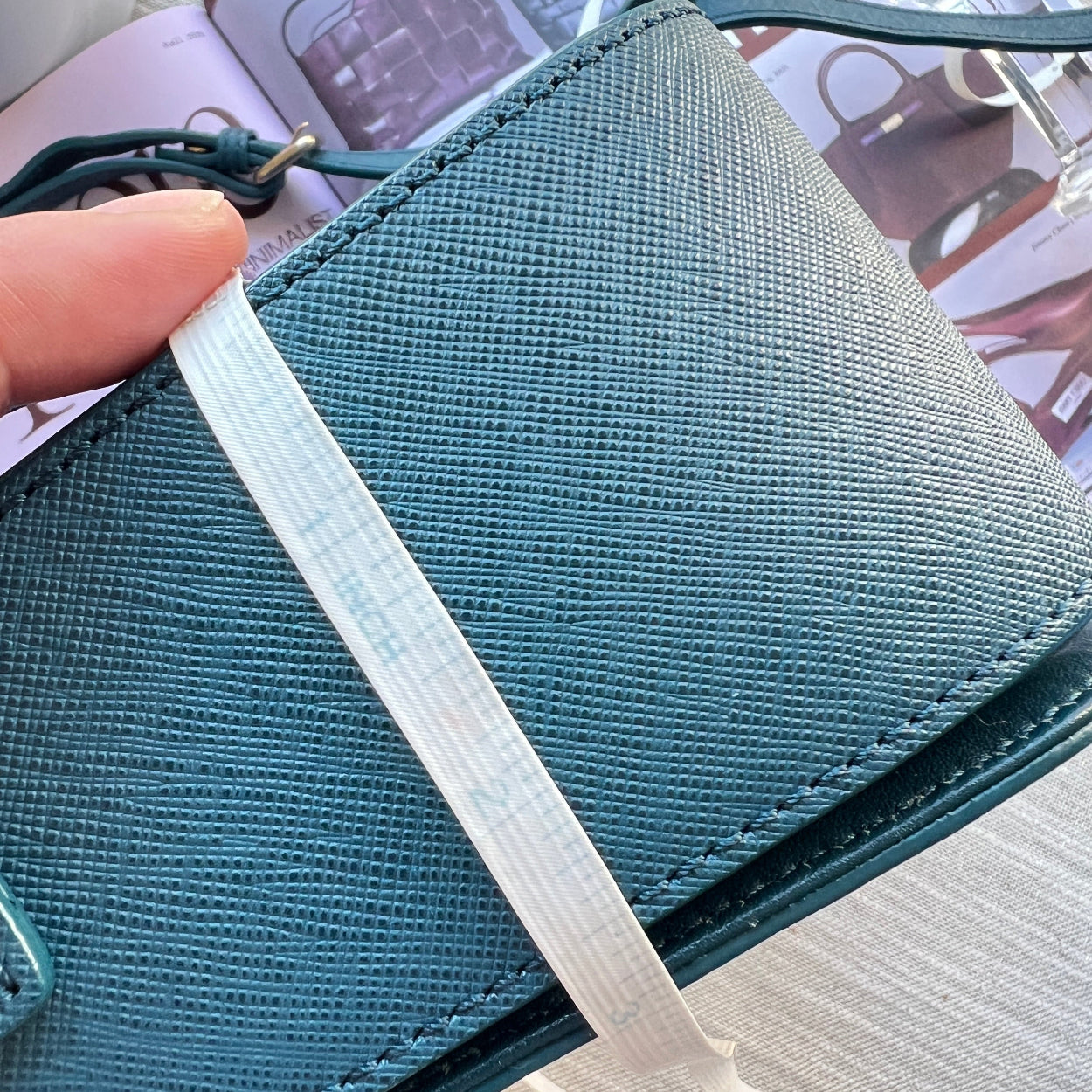 Teal fabric with a white strap held by a hand, with a magazine in the background.