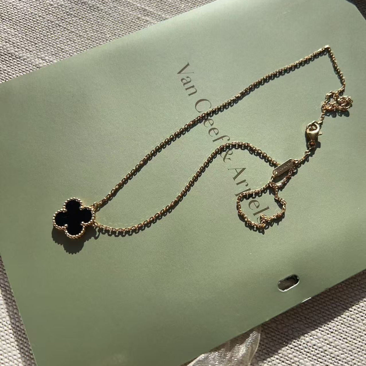 A gold-tone Van Cleef & Arpels necklace with a black clover pendant and a heart-shaped charm, displayed on its light green branded presentation card.