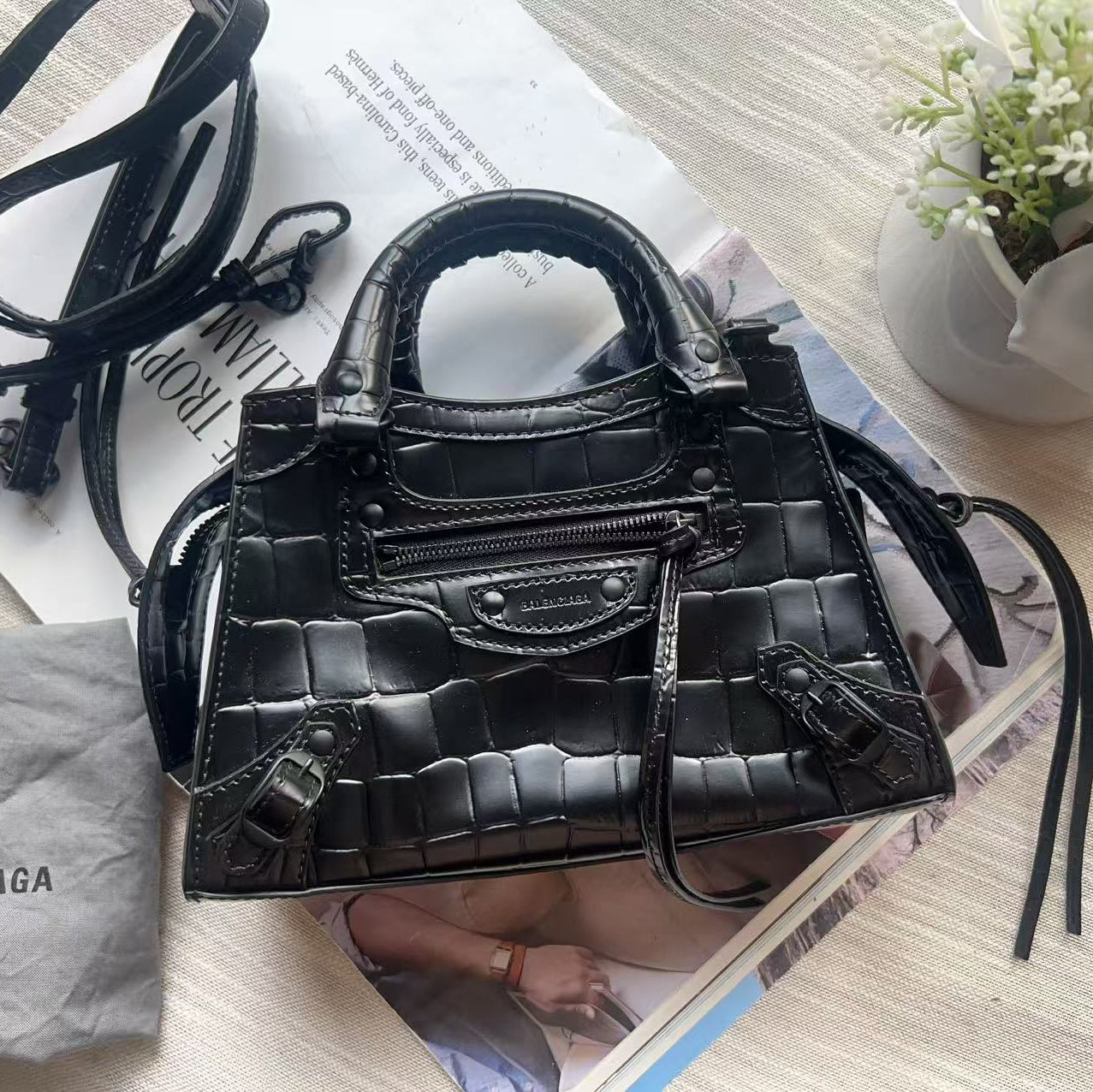 A black Balenciaga crocodile-embossed leather handbag with top handles and a detachable shoulder strap, styled on top of a magazine with a gray Balenciaga dust bag and white flowers in the background.