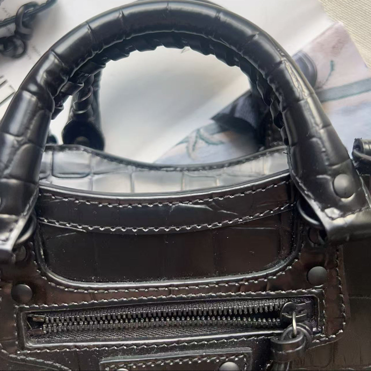 Close-up of a dark brown crocodile-embossed leather handbag showing the handle attachment, zipper, and stitching details.