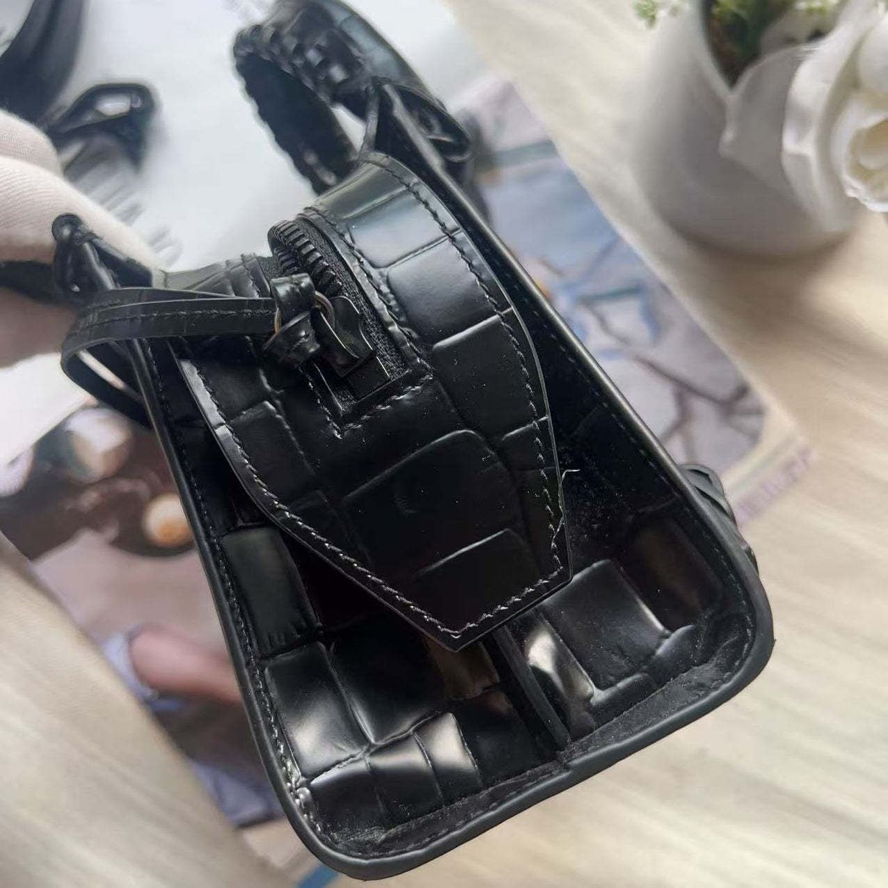 A close-up of a black crocodile-embossed leather handbag, showing the interior lining, zipper closure, and strap attachment, resting on a light-colored wooden surface with a magazine and flowers in the background.