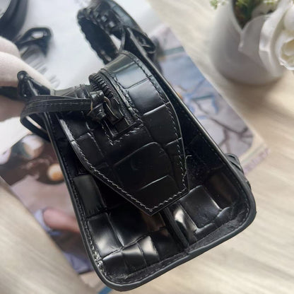 A close-up of a black crocodile-embossed leather handbag, showing the interior lining, zipper closure, and strap attachment, resting on a light-colored wooden surface with a magazine and flowers in the background.