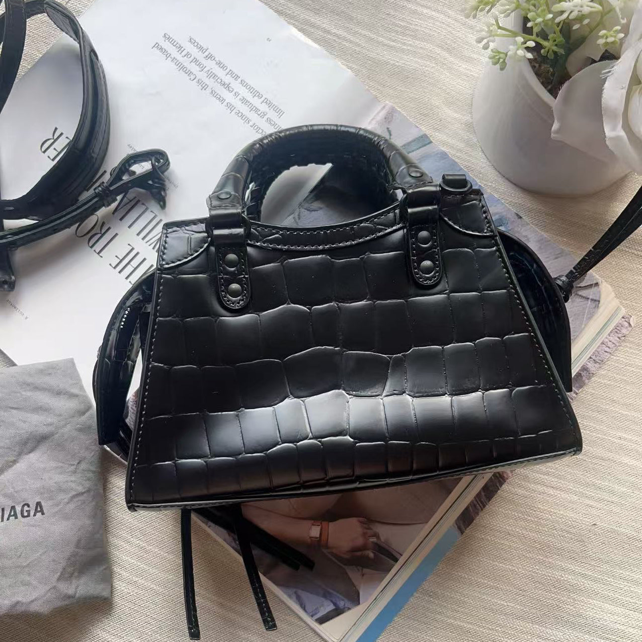 A black crocodile-embossed leather handbag with top handles and a shoulder strap, styled on a light beige fabric surface with printed papers, a gray dust bag, and a small floral arrangement.