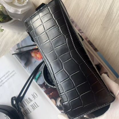A black crocodile-embossed leather clutch or wallet-style handbag held by a white gloved hand, resting on top of a magazine against a light beige fabric background.