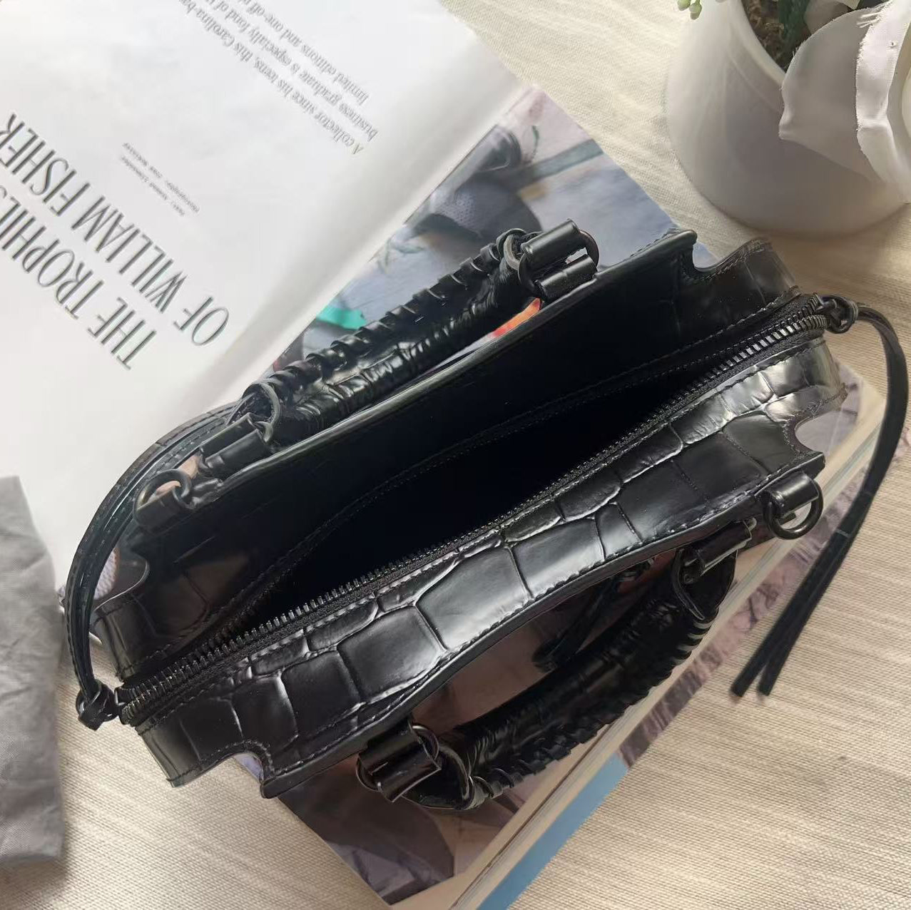 A black crocodile-embossed leather handbag with braided details, shown open on a light wood tabletop with an open magazine underneath. A gray Balenciaga dust bag is partially visible on the left.