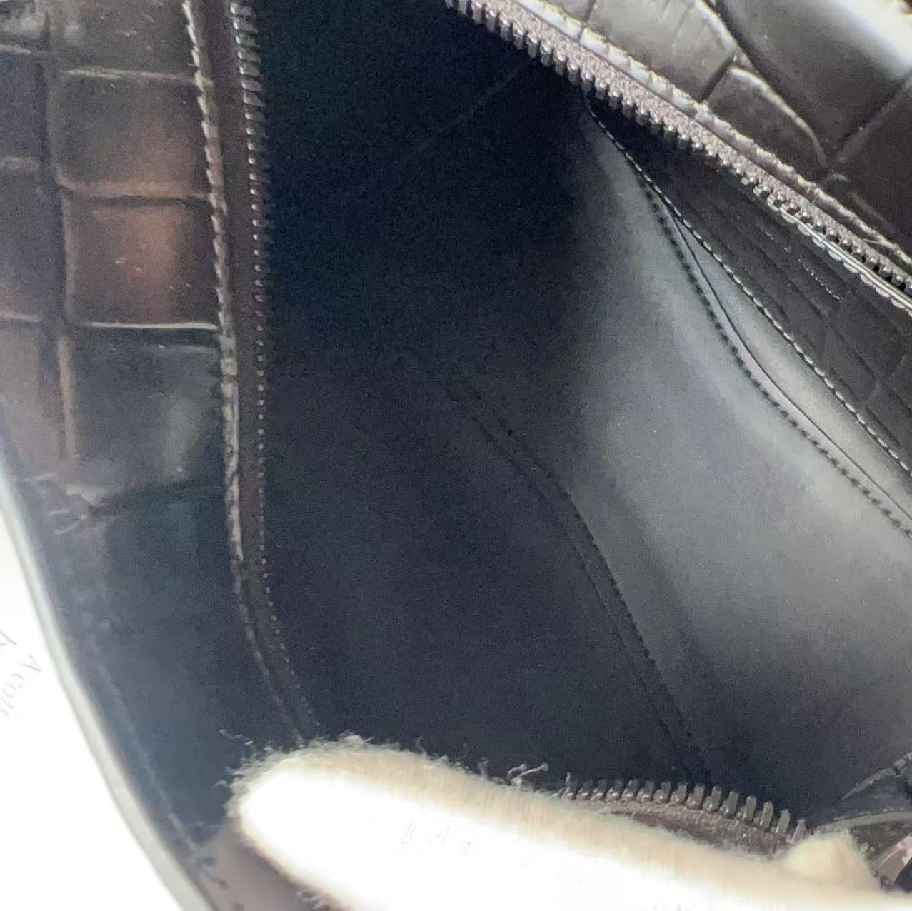 Close-up of the interior of a black crocodile-embossed leather bag, showing the black fabric lining and zipper, with a white gloved hand holding the opening.