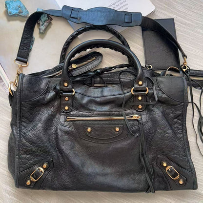 A black leather shoulder bag with gold-tone hardware, laid flat on a light wood surface next to a white paper and small blue stones.