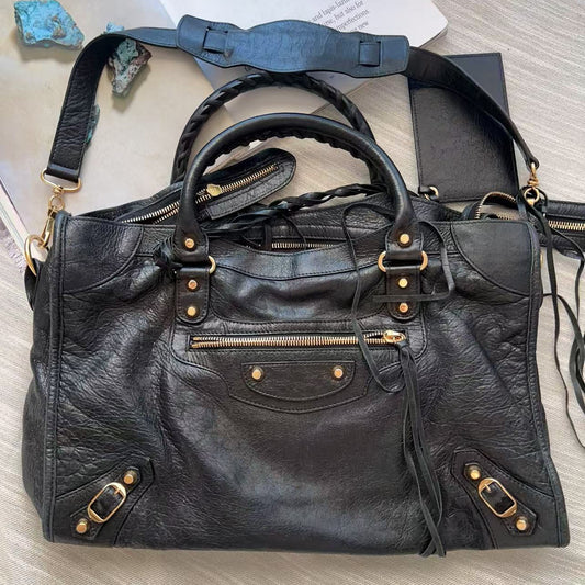 A black leather shoulder bag with gold-tone hardware, laid flat on a light wood surface next to a white paper and small blue stones.