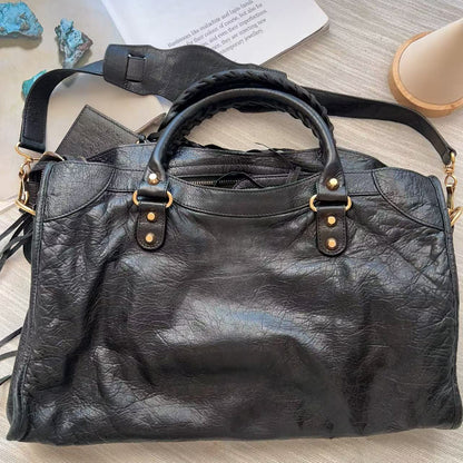 A large black leather handbag with distressed texture and gold-tone hardware, laid flat on a light wood surface next to a book and small stones.