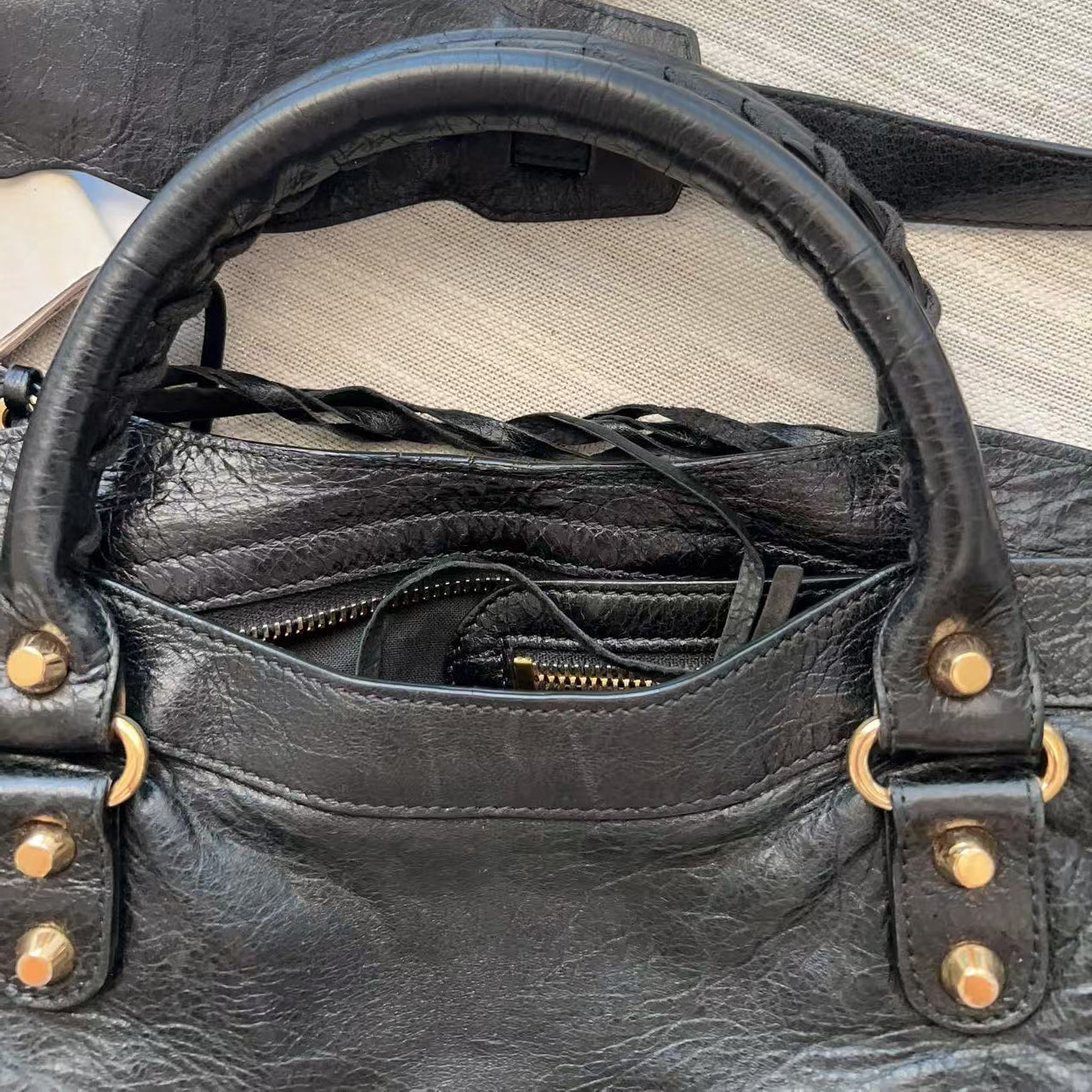 A close-up of a black distressed leather handbag showing the interior zippered compartment, braided handle details, and brass-tone hardware on a neutral textured surface.