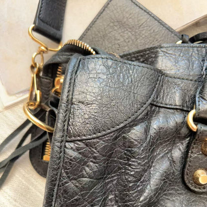 Close-up of a black pebbled leather handbag showing the texture, gold-tone hardware, and part of the strap on a light neutral surface.