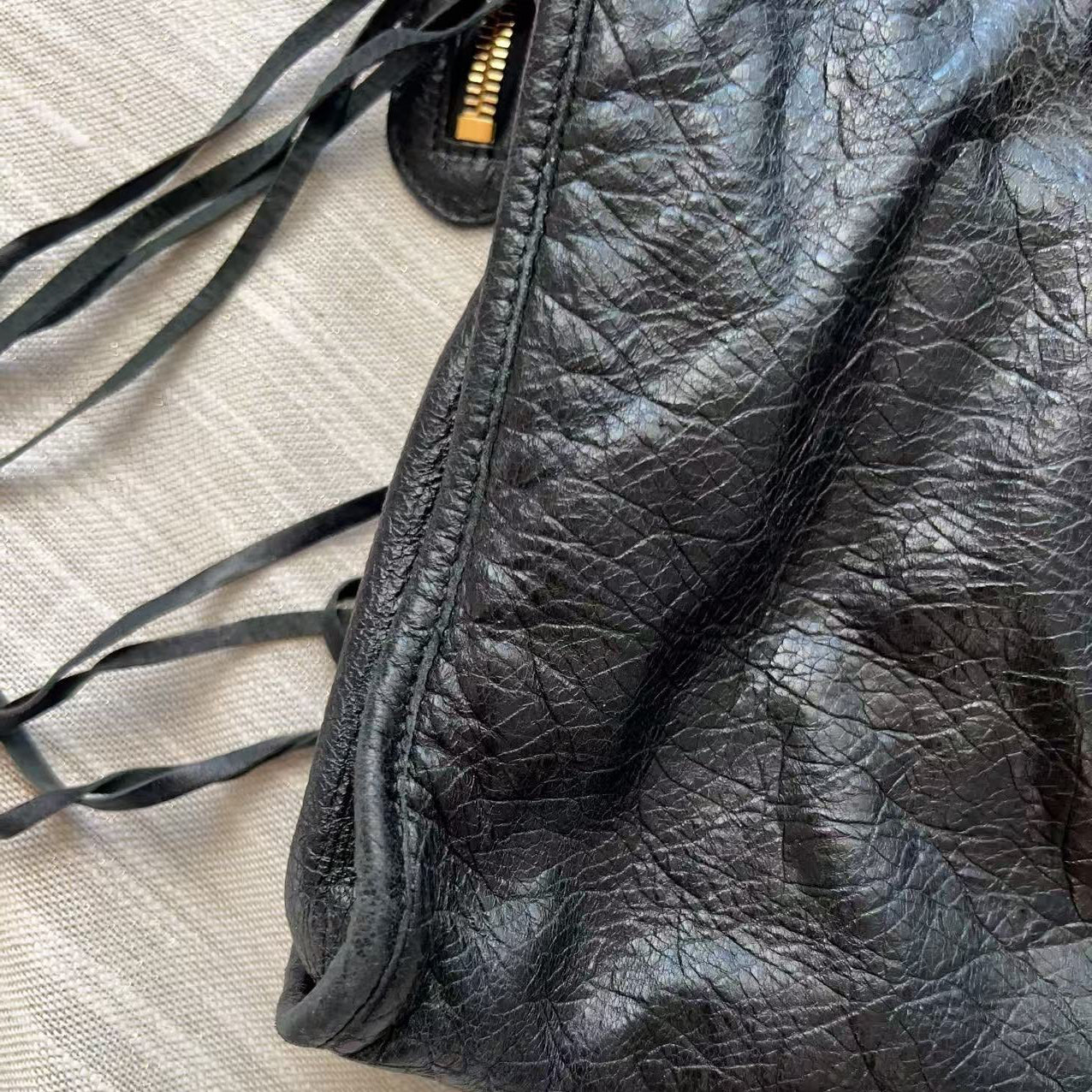 Close-up of a black crinkled leather bag corner with a gold zipper pull and dark drawstring ties, placed on a light beige textured fabric surface.