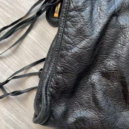 Close-up of a black crinkled leather bag corner with a gold zipper pull and dark drawstring ties, placed on a light beige textured fabric surface.