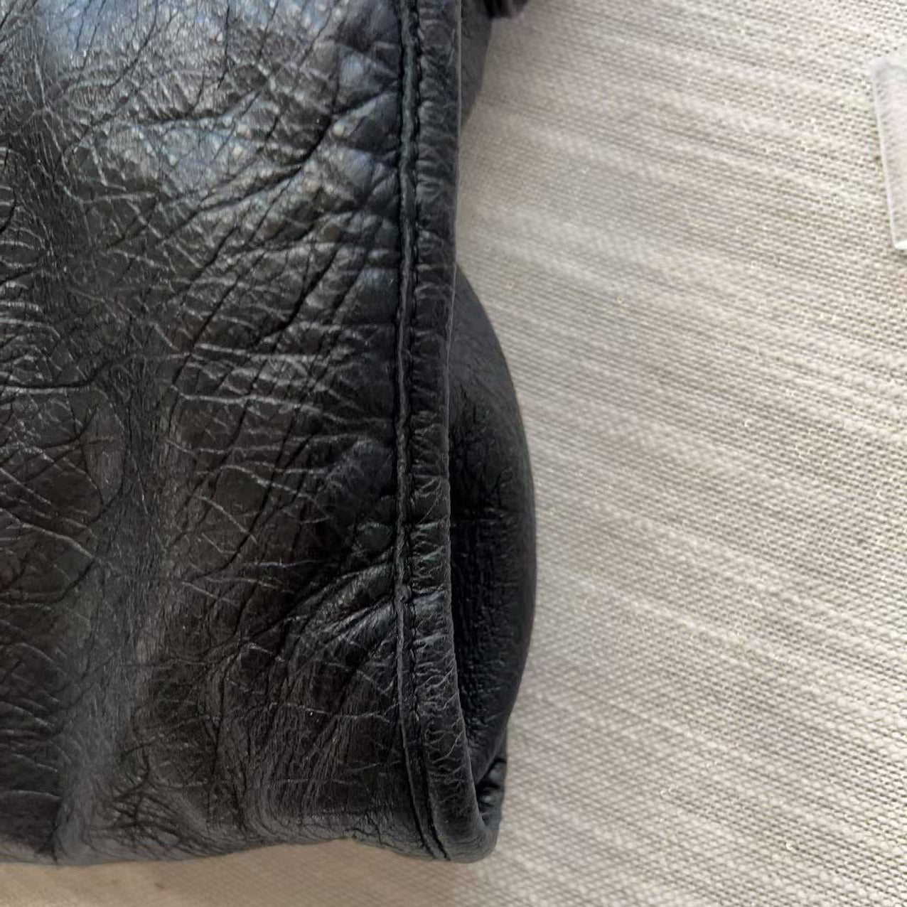 A close-up of a black leather bag corner showing textured leather grain and a stitched rim, set against a light beige woven fabric background.