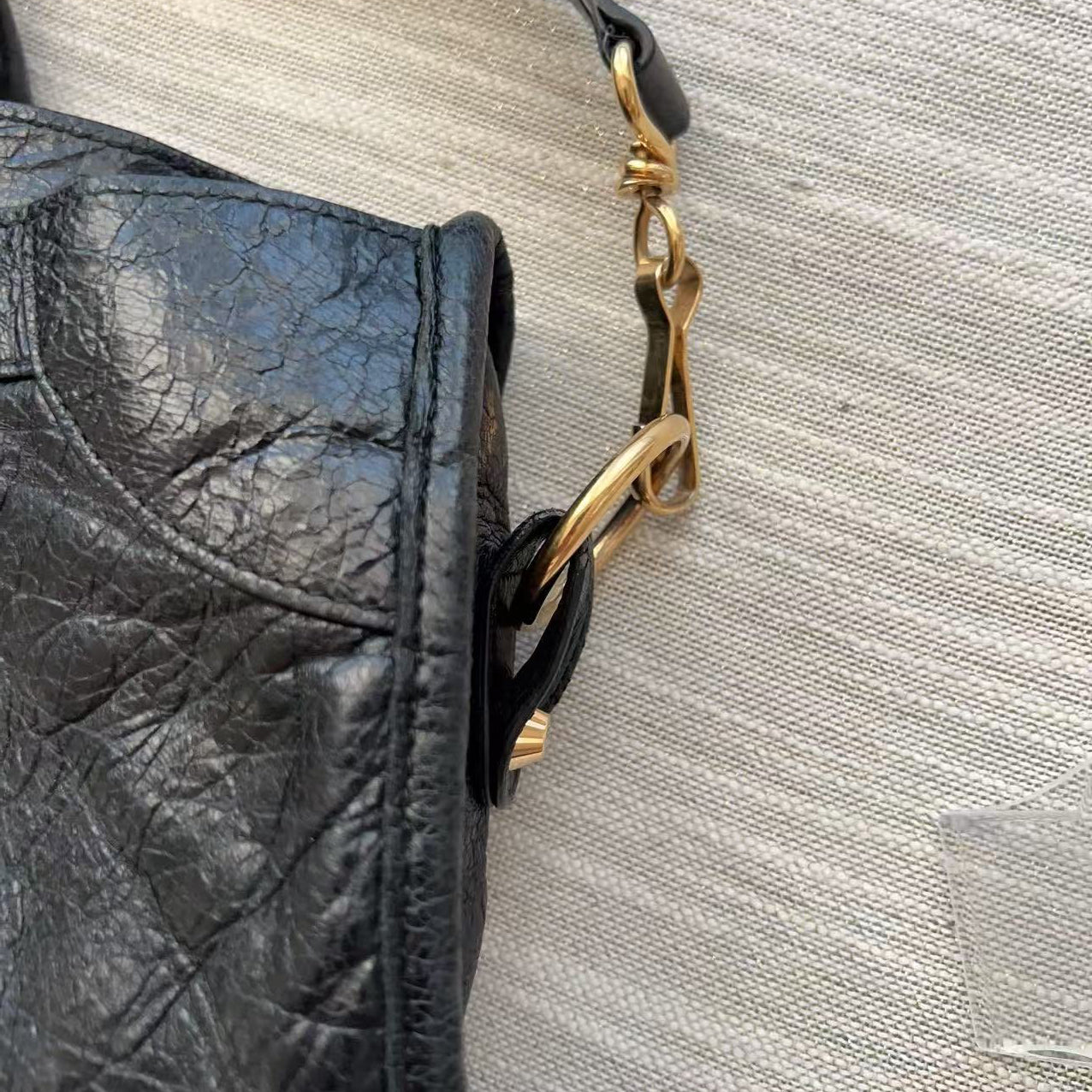 Close-up of a black textured leather handbag with gold-tone hardware and a black strap against a light beige woven fabric background.