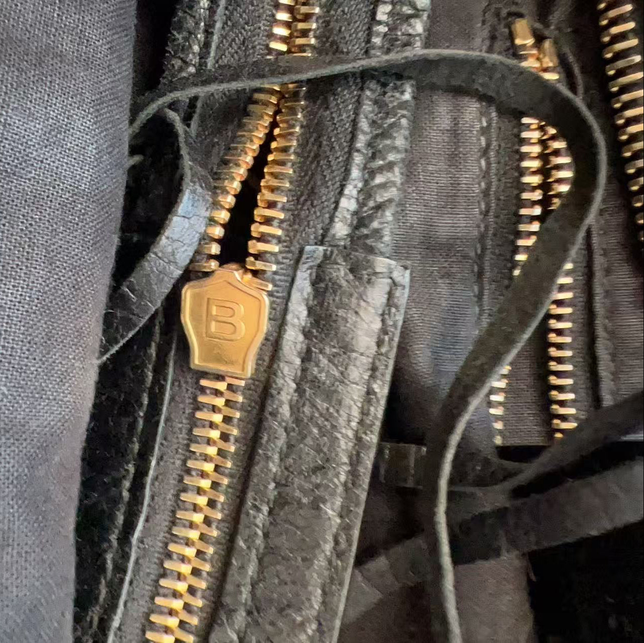 Close-up of a dark gray fabric bag with multiple gold-tone metal zippers and a small gold zipper pull featuring an embossed letter B.