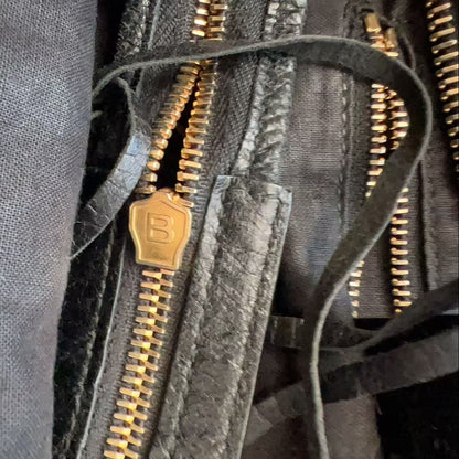 Close-up of a dark gray fabric bag with multiple gold-tone metal zippers and a small gold zipper pull featuring an embossed letter B.
