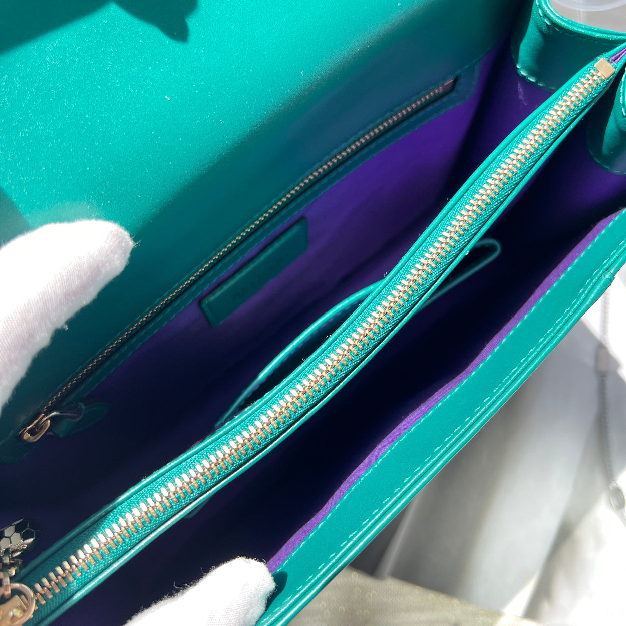 Close-up of a teal handbag with a gold zipper, held by a white gloved hand.