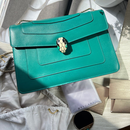 Teal handbag with decorative clasp on a surface with a magazine and flowers.