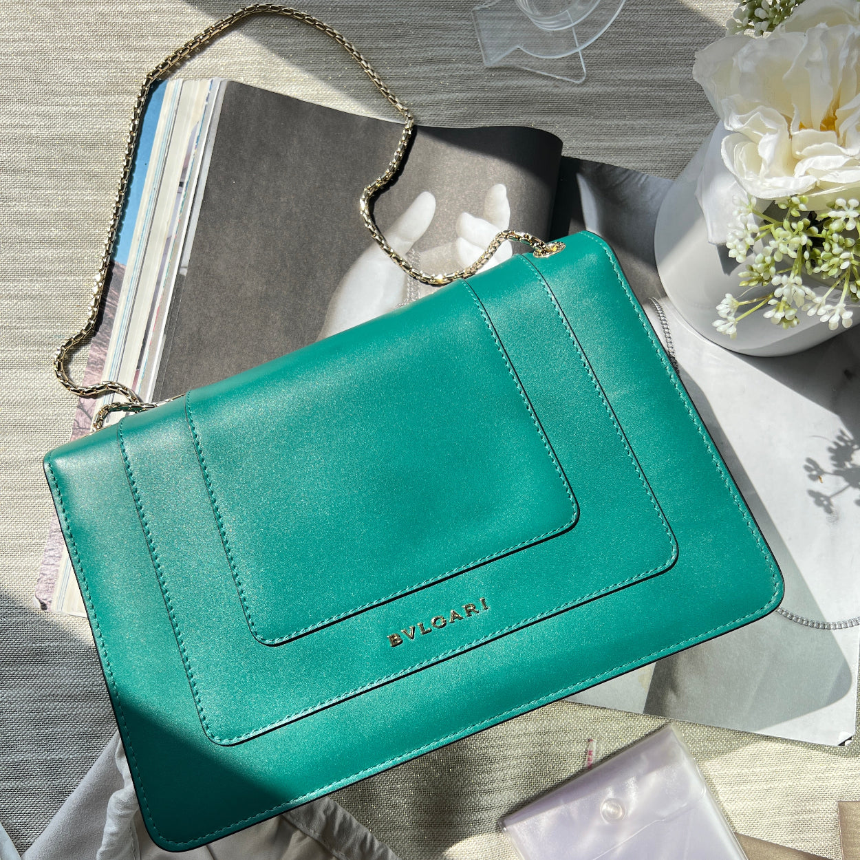 Teal handbag with chain detail on a surface with books and flowers