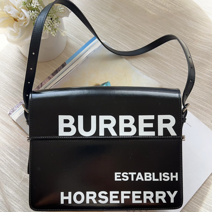 Black handbag with 'BURBER' and 'HORSEFERRY' text on a light wooden surface.
