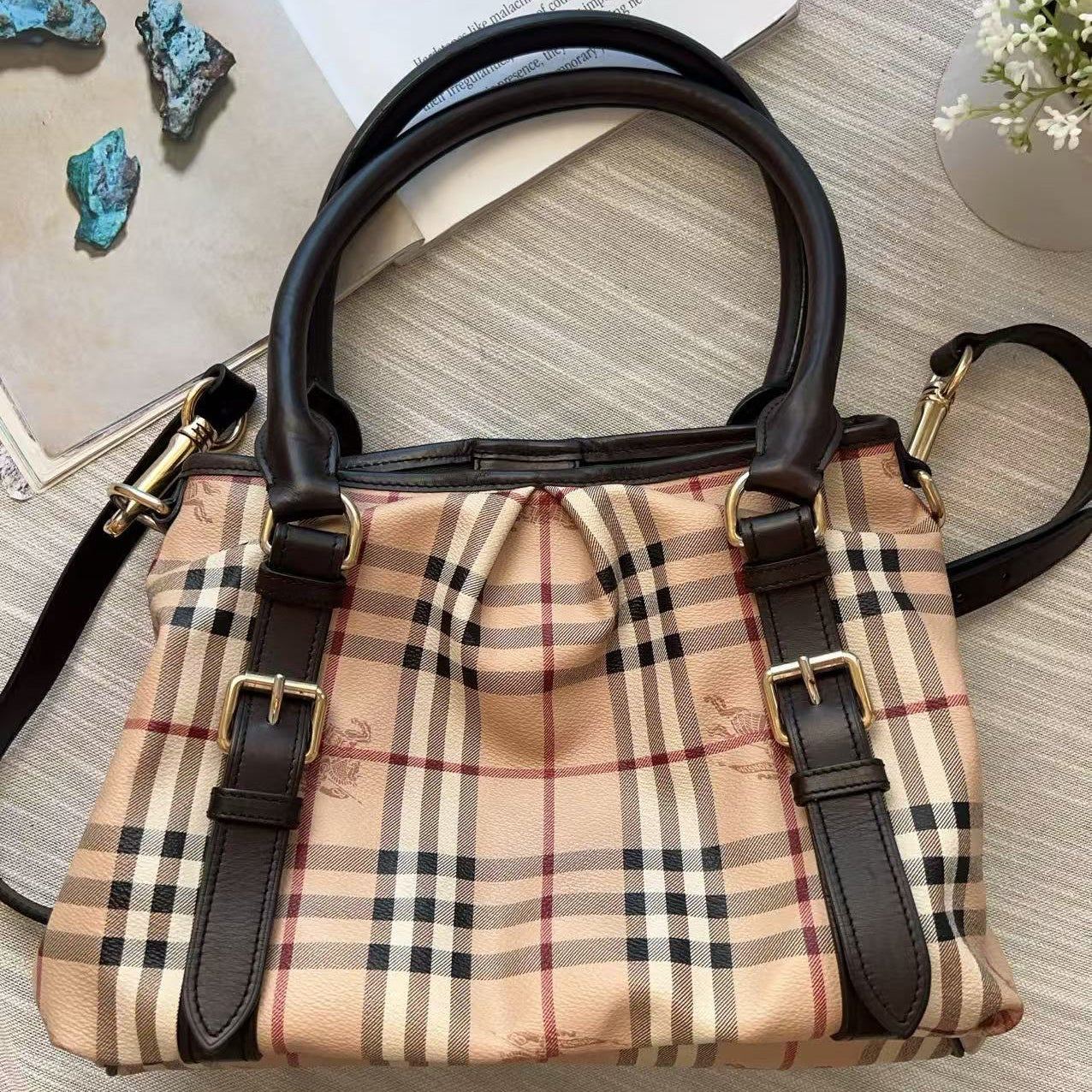 A beige plaid handbag with black leather trim and gold-tone hardware, photographed from above on a light wood surface with a few styled props nearby.