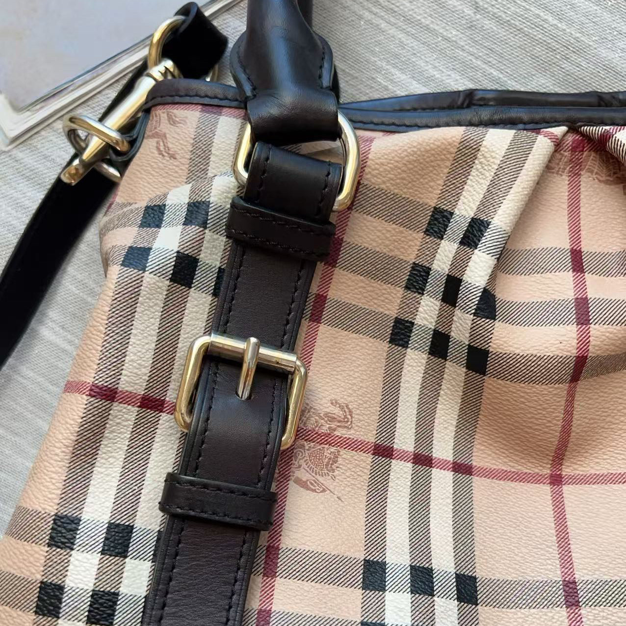 Close-up of a beige plaid handbag with black leather trim and gold-tone hardware, showing part of the strap and buckle details.
