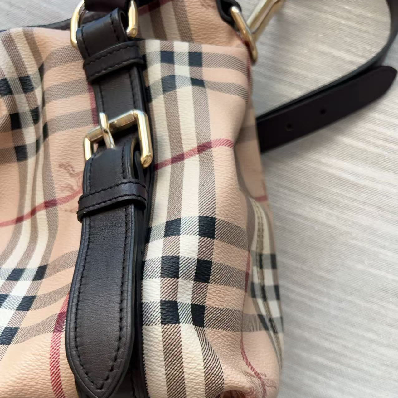 Close-up of a beige plaid handbag with black leather strap and gold-tone buckle, resting on a light gray textured surface.
