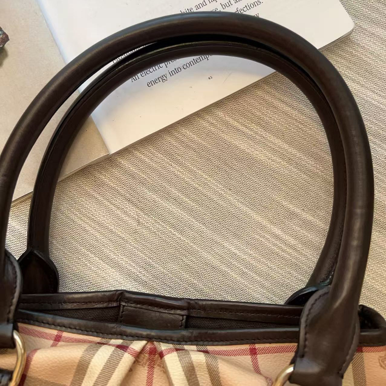 A close-up of a handbag with dark brown leather handles and a beige plaid pattern, resting on a textured beige surface next to a white book or magazine.