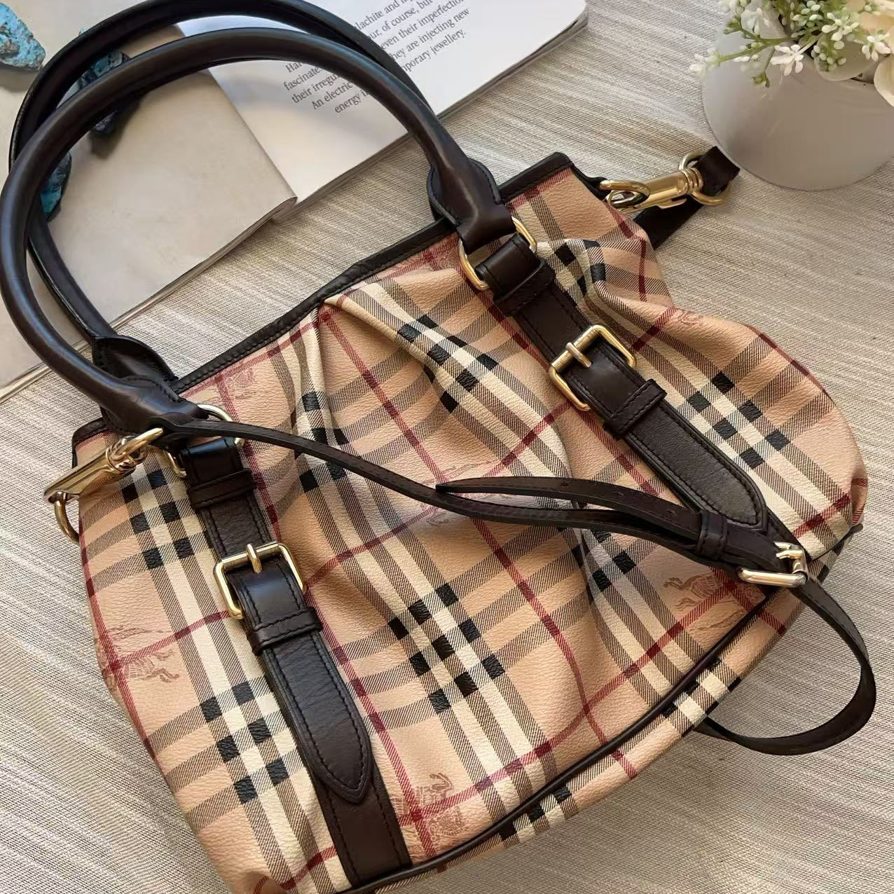A classic Burberry-style plaid handbag in beige, black, and brown, featuring dark brown leather trim, double top handles, and gold-tone hardware, laid flat on a light wood-textured surface.