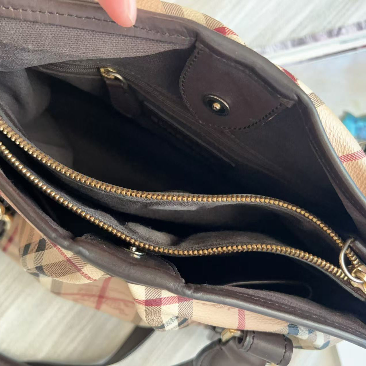 Close-up of the interior of a plaid handbag with dark brown leather lining, gold-tone zippers, and a beige plaid pattern visible on the outer fabric.