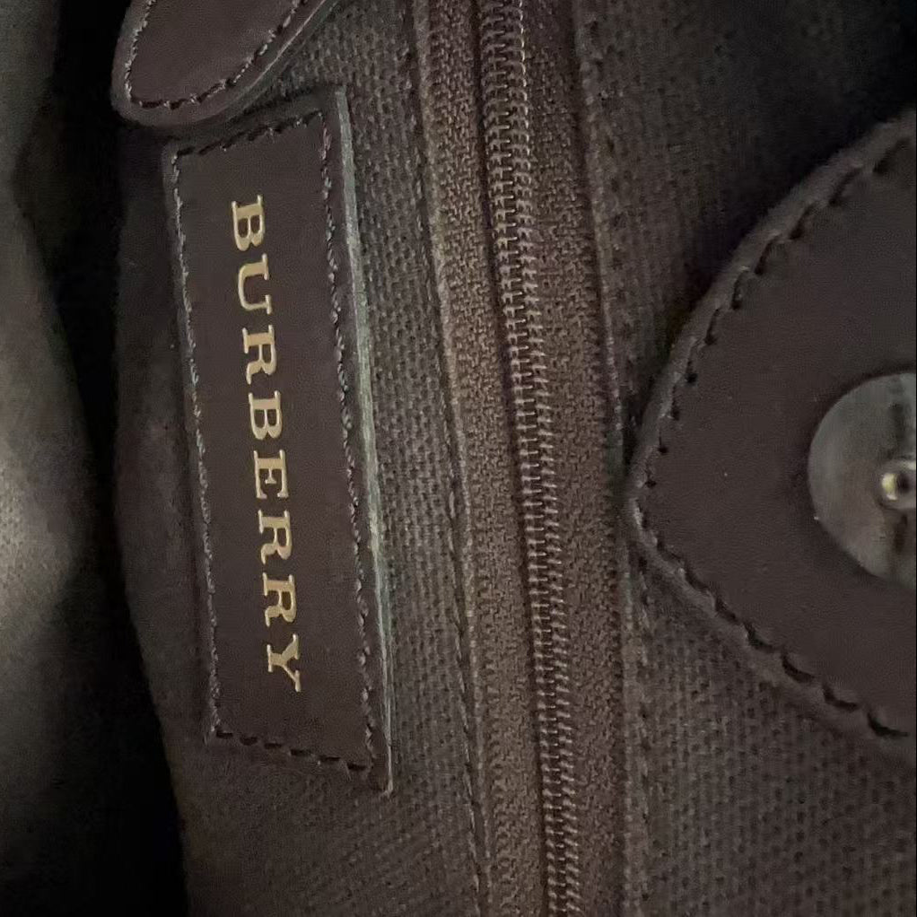 Close-up of a dark brown textured canvas bag with a brown leather patch displaying the gold Burberry logo next to a zipper.