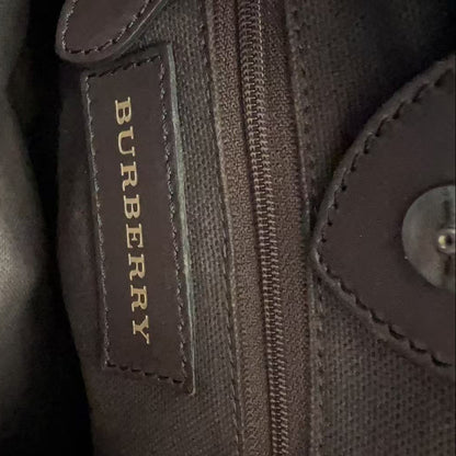 Close-up of a dark brown textured canvas bag with a brown leather patch displaying the gold Burberry logo next to a zipper.