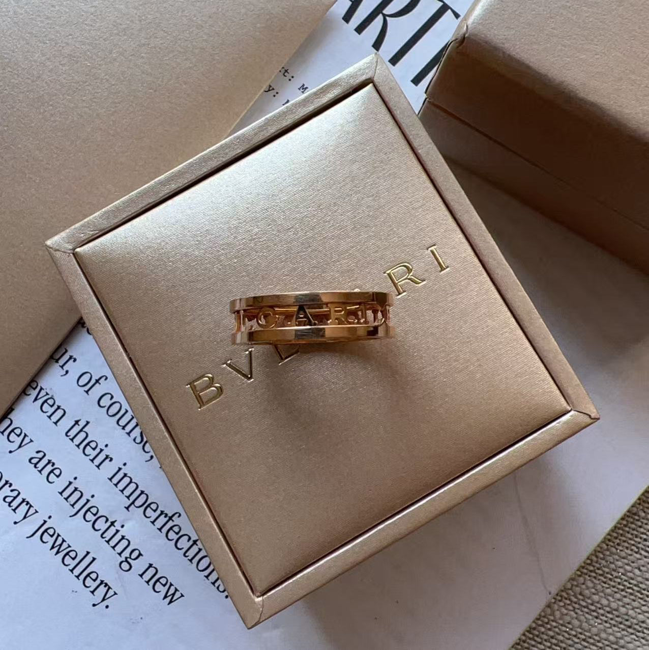 A gold ring with a central red inlay, displayed on a beige Bvlgari box lid. The box is placed on top of a book page with the brand name 'BVLGARI' partially visible, alongside other matching beige packaging boxes.
