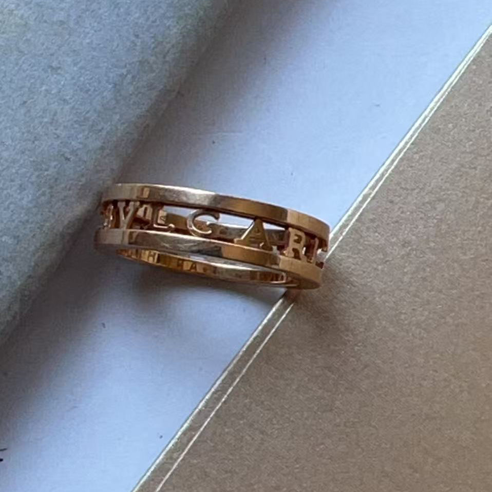 A gold ring with the brand name 'BVLGARI' engraved across it, resting on a light blue textured surface next to a brown paper card.