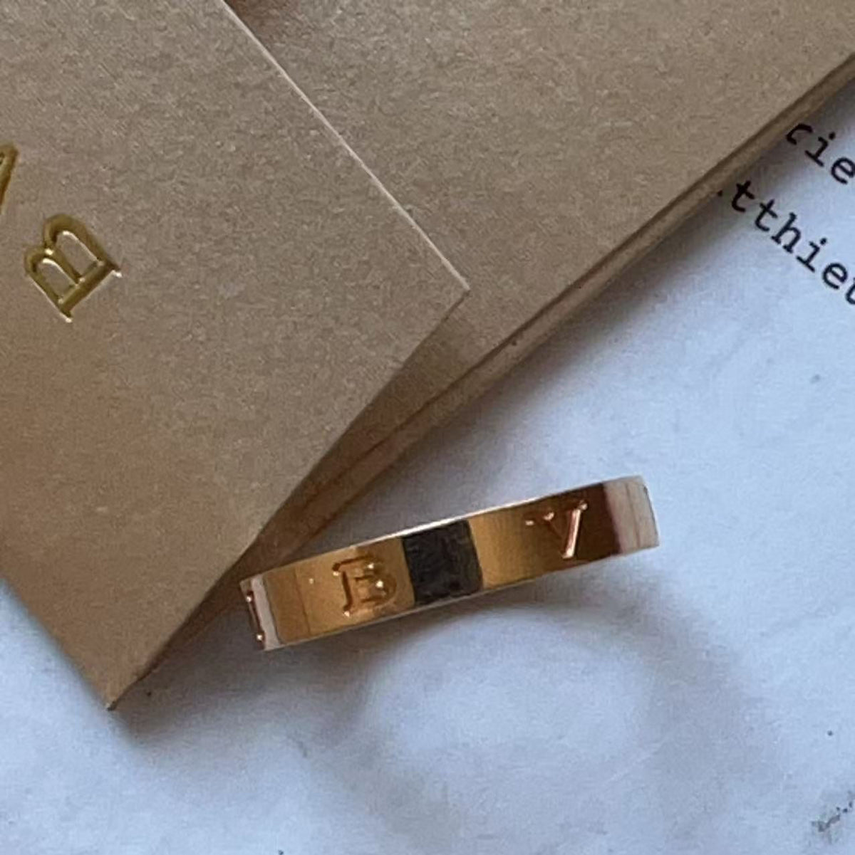 A gold-toned ring with engraved letters 'E' and 'Y' and a central black inlay, resting on a light surface next to brown kraft paper packaging.
