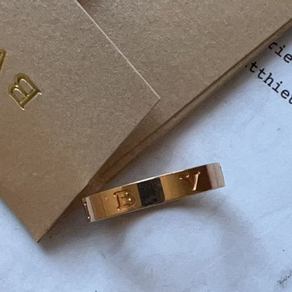 A gold-toned ring with engraved letters 'E' and 'Y' and a central black inlay, resting on a light surface next to brown kraft paper packaging.
