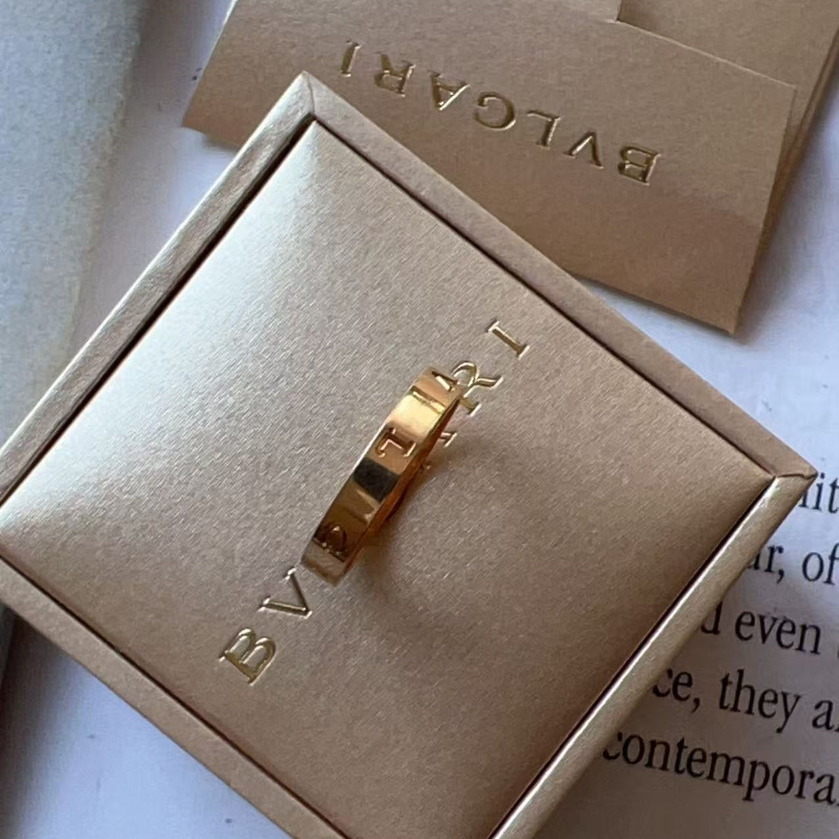 A gold ring with interlocking square-cut stones, presented in a gold-tone jewelry box embossed with 'BVLGARI', with its branded card behind it on a light surface.